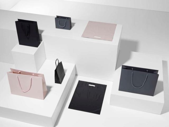 Esposizione di borse per regali in vari colori e dimensioni, su diverse piattaforme bianche. Le borse includono opzioni nere e rosa con manici, ideali per confezioni regalo eleganti.